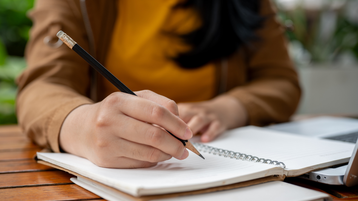A close-up image of an Asian woman holding a pencil, writing something in her notebook, making list, drawing, taking notes, doing homework, or keeping diary. freelance writing jobs.