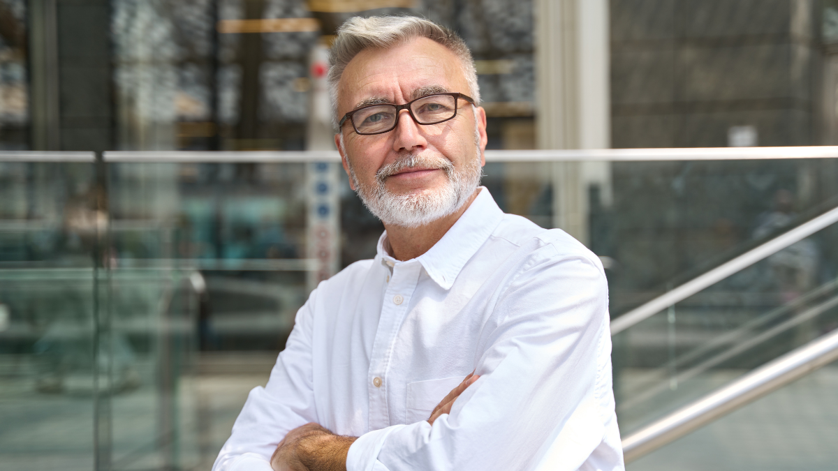 Confident happy mature older business man leader, smiling middle aged senior old professional businessman wearing white shirt glasses crossed arms looking at camera standing outside, portrait.