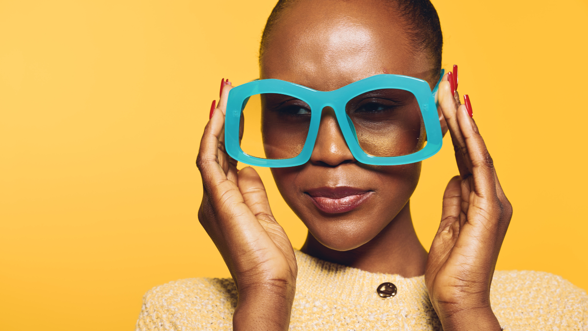 Cheap Prescription Glasses Close-up of a confident dark skinned African woman wearing oversized blue glasses and a beige sweater against a bright yellow background. Concept of fashion, style, and beauty.