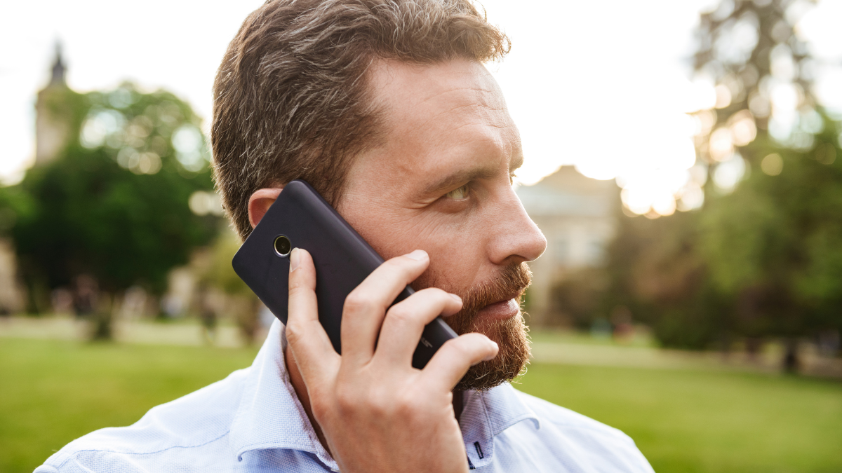 Portrait closeup of serious man 40s in white shirt looking aside during walk in green park while talking on black smartphone risk averse.