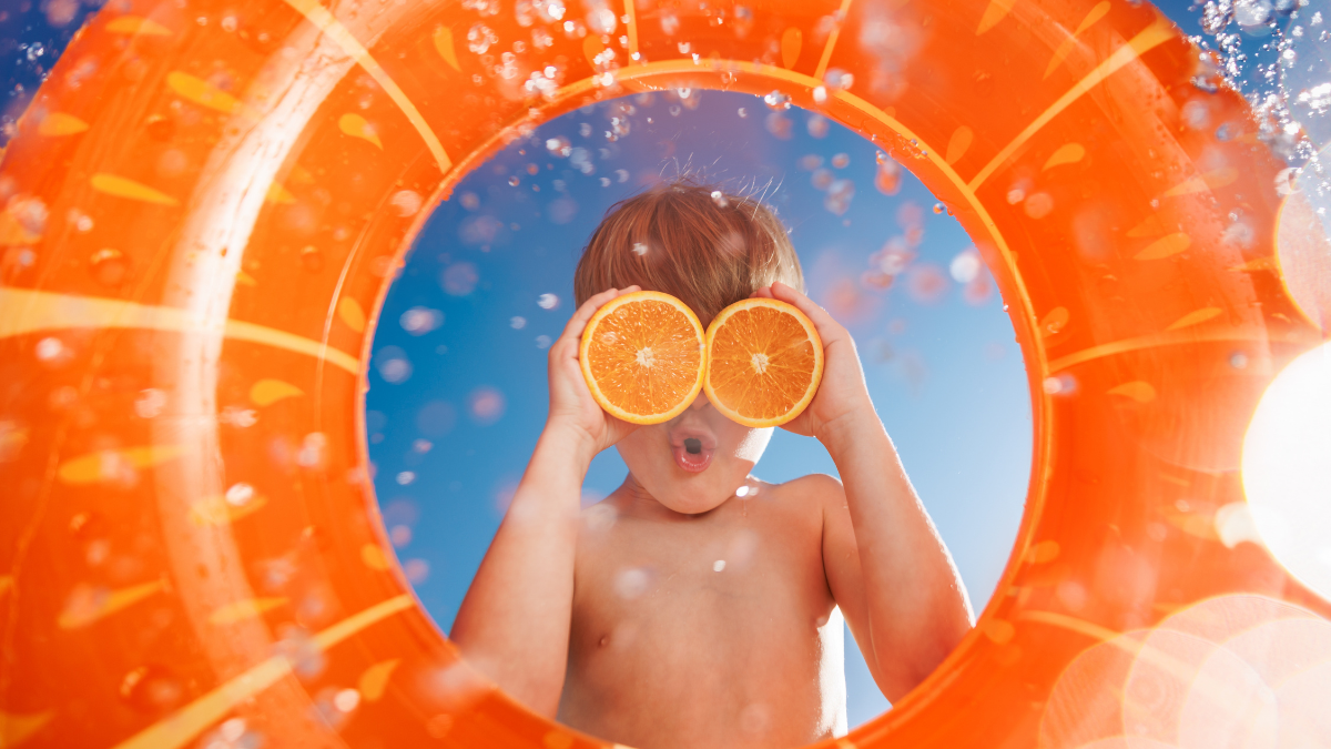Happy child holds the orange halves like sunglasses. Kid having fun on summer vacation. Boy jumping into swimming pool with splashes of water. Funny child against blue sky background.