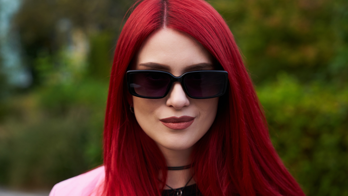 Young woman with vibrant red hair wears denim jacket in a city park.
