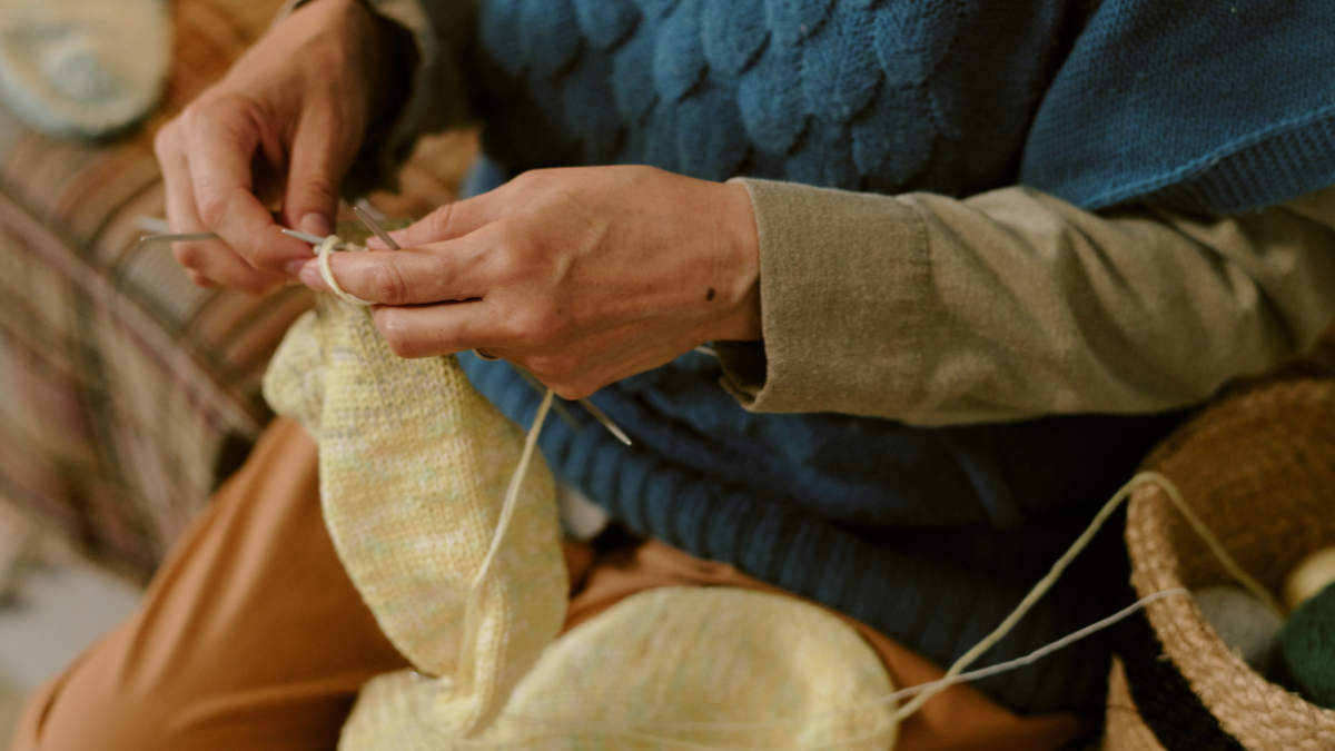 Senior Caucasian woman knitting with yarn, hands holding knitting needles while creating fabric, sitting indoors, focusing on craft, partial view showing detailed hand movement frugal living.