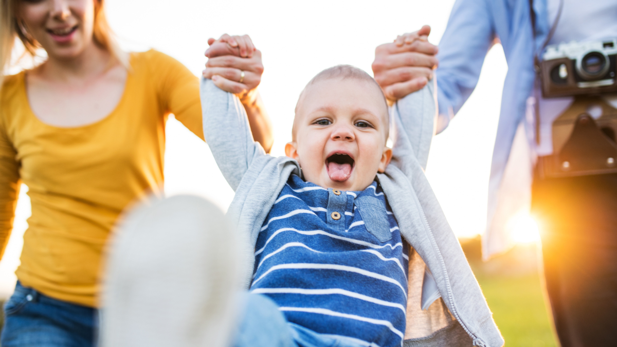 New parents holding small toddler, by hands, swinging him. Family outdoors on walk in spring nature. Happy family moment.