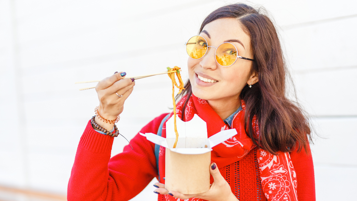 Bright hipster woman eating asian fast food from takeaway box with chop sticks, Wok noodles concept cheap dinner ideas.