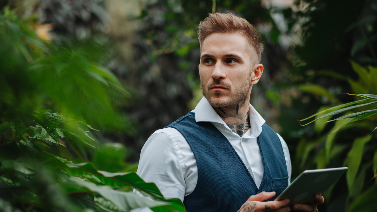 Handsome businessman with tablet, standing in botanical garden, in the middle of lush green foliage. Concept of business corporate social responsibility and ESG Impact Investing.