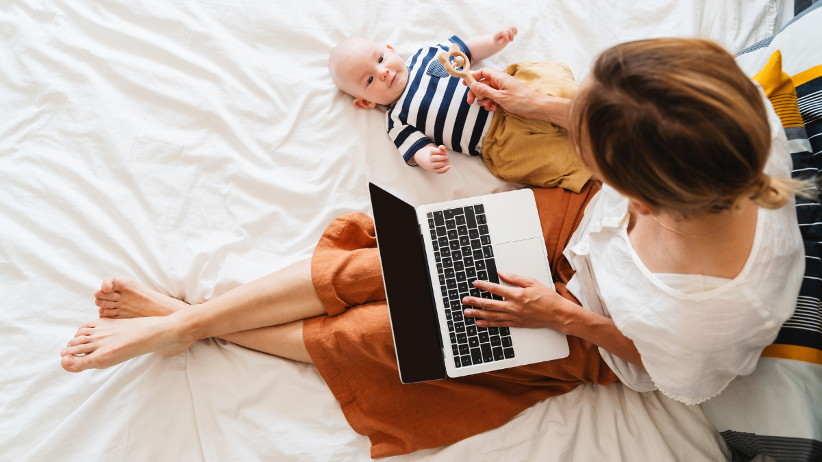 Mother working at laptop playing with baby in bed at home. Modern woman balances between work and baby. Child playing with toy on bed while mother using laptop: surfing internet, doing online shopping.