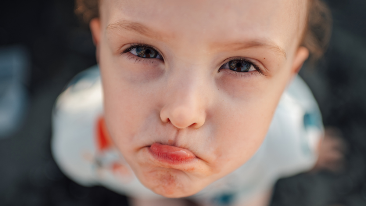 Headshot of a cute little boy with sad face looking at the camera. Close up of an unhappy kid sitting in sand on a beach on summer vacation. From above view of a distressed kid looking at the camera. How To Save Money for Kids SH