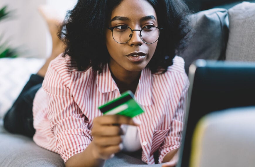 Skilled African American female freelancer installing paid application for doing web designing checking data debit info for making online commerce payment, woman using credit card for online banking.