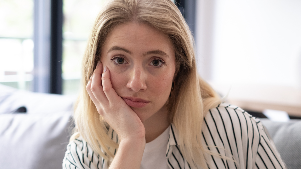 A pensive young woman sits on a sofa at home with her face resting on her hand, staring thoughtfully. Natural light and a striped shirt create a quiet, contemplative, slightly bored mood. Frugality vs. Cheapskate.