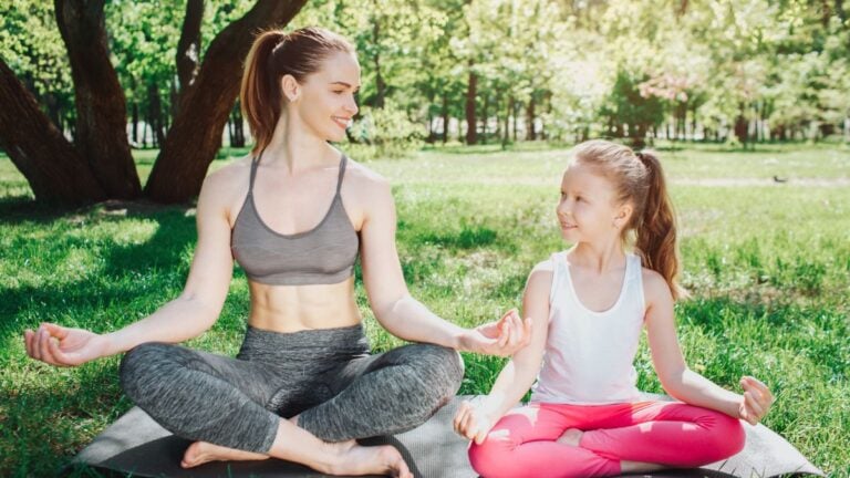 mother and daughter are sitting on carimate in lotus pose and loking at each other