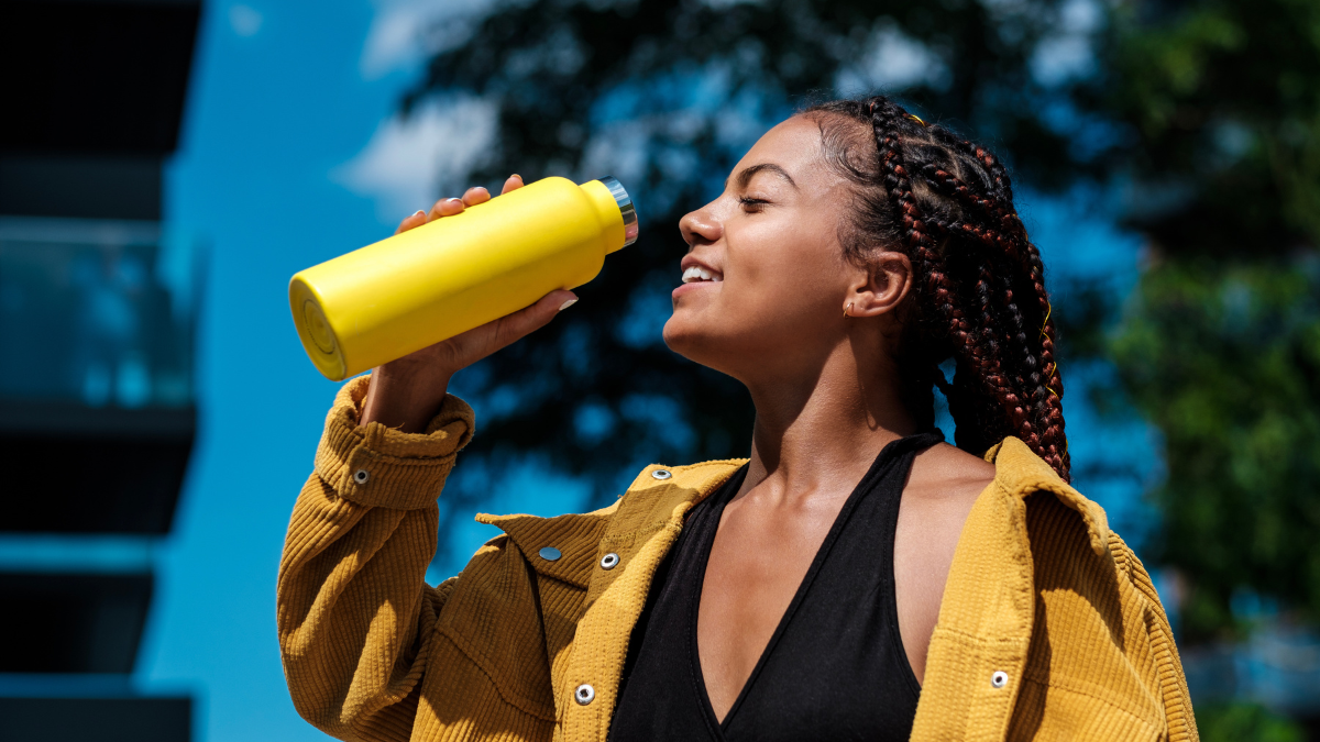 Attractive young woman drinking from a yellow bottle. She is wearing a yellow jacket and it is a hot day in summer. Smiling. Mixed race. frugal purchases woman with thermos.