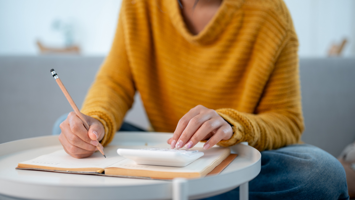 A close-up image of a woman sitting on a sofa at home, writing in a book and using a calculator to manage her household expenses and calculate her monthly savings. people and financial concepts,