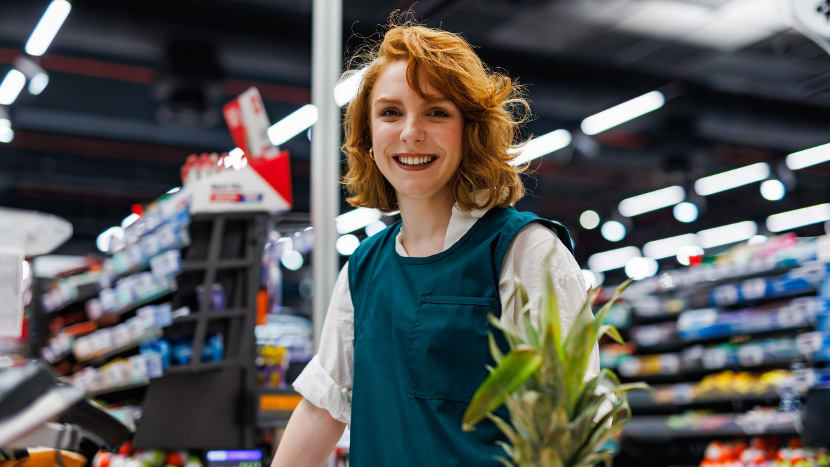 Happy young supermarket employee wearing uniform holding pineapple while working at checkout counter, smiling at camera in grocery store.