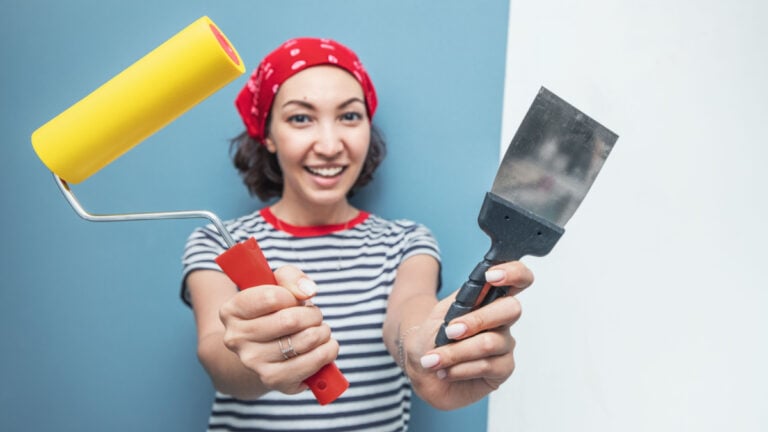 Happy woman repairman poses with a roller and trowel against the background of pasted wallpaper and a plastered wall.