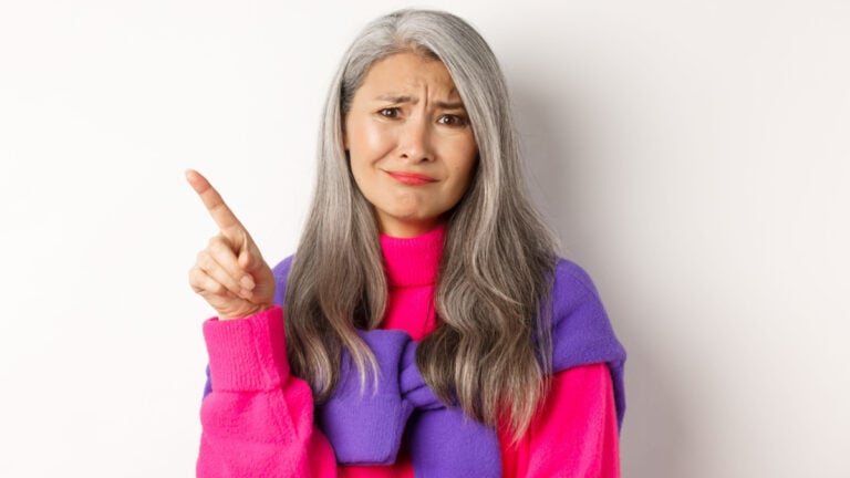 Judgy and skeptical asian senior woman grimacing displeased, pointing finger upper left corner unamused, standing over white background.