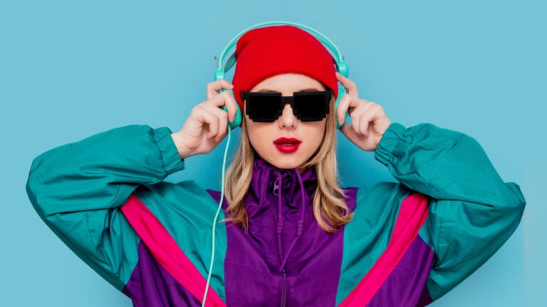 Portrait of a woman in red hat, sunglasses and suit of 90s with headphones on blue background./