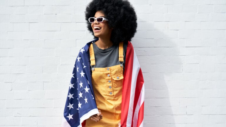Happy young African American woman wrapped in USA flag laughing standing against white wall.
