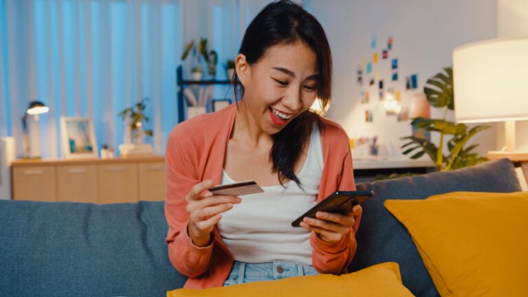 Night time beautiful smiling asian lady use cellphone order online shopping product with credit card on sofa in living room.