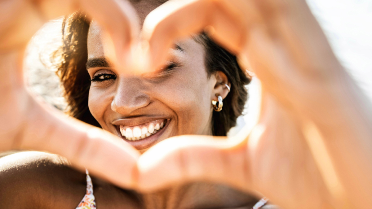 Close up image of smiling woman in swimwear on the beach making a heart shape with hands.