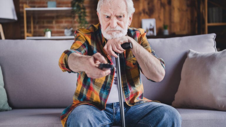Photo of angry irritated man pensioner wear checkered shirt changing tv channels holding walking cane sitting sofa indoors house room.