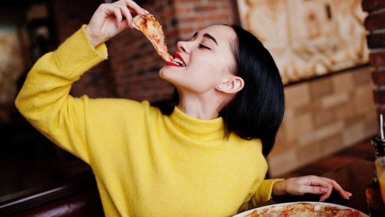 Funny brunette girl in yellow sweater eating pizza at restaurant..