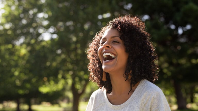Young woman with curly hair laughing while standing outside in a park on a sunny summer afternoon.