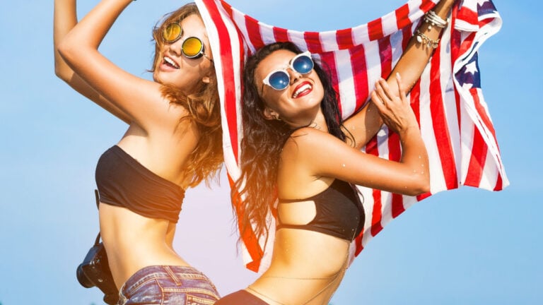 Two beautiful tanned fun hipster girls on the beach, blonde and brunette holding an American flag.