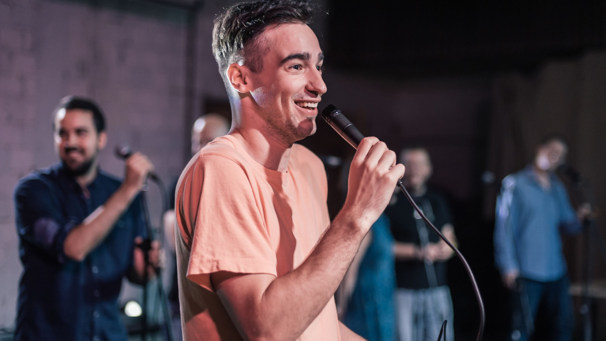 Smiling and witty stand up actor comedian on stage, talking and singing into a microphone and making the audience laugh.