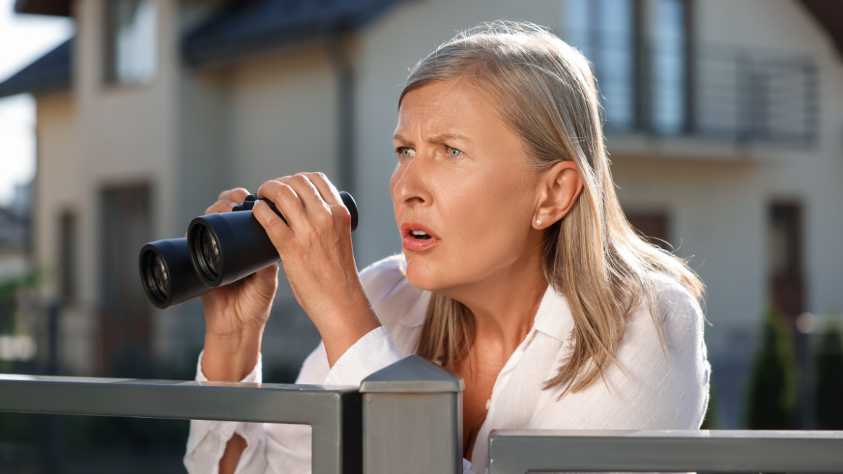 Concept of private life. Curious senior woman with binoculars spying on neighbours over fence outdoors.