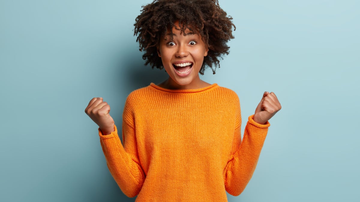 Joyful dark skinned model with crisp hair, clenches fists, feels overjoyed after winning game, wears orange jumper, poses over blue background, expresses good emotions.