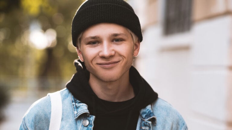 Smiling teen boy 16-18 year old wearing black knitted hat, hoodie and denim jacket outdoors at sun set on background. Looking at camera. Teenagerhood.