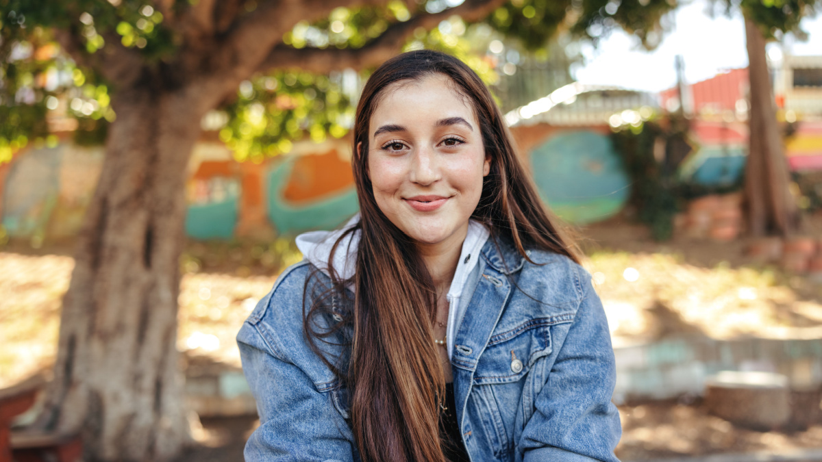 Happy teenage girl looking at the camera with a smile. Cheerful brunette girl wearing a denim jacket outdoors in the city. Female youngster sitting alone in an urban park during the day.