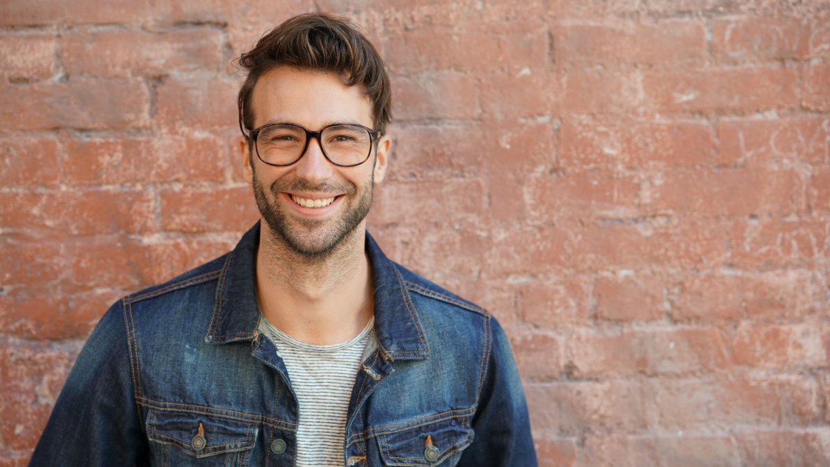 Smiling trendy guy with blue jeans jacket and eyeglasses standing by brick wall.