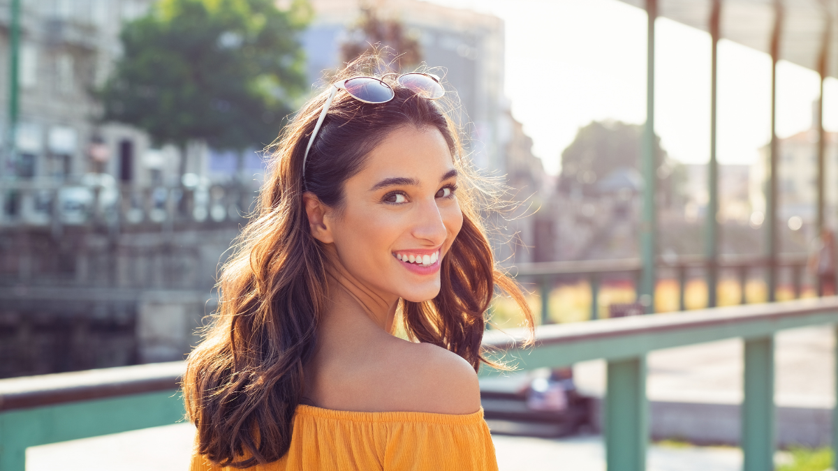 Happy latin woman looking behind while walking on street. Beautiful young woman enjoying vacation during summer. Cheerful girl in smart casual walking and looking at camera while smiling.