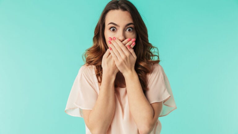 Portrait of a shocked young girl in dress looking at camera with mouth covered isolated over blue background.