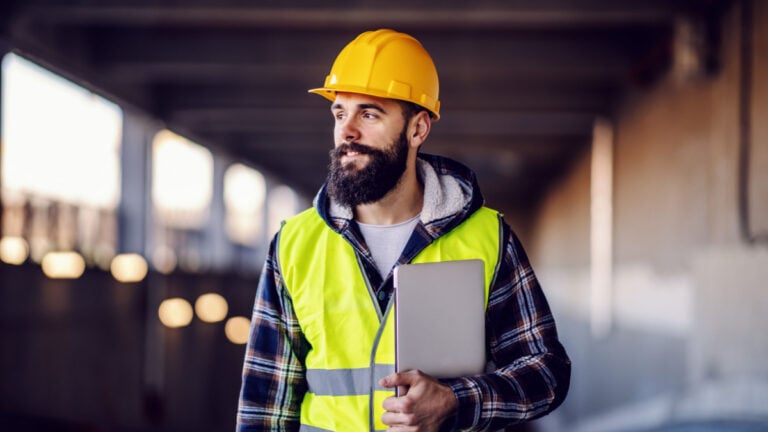 Portrait of highly motivated caucasian hardworking smiling bearded supervisor with helmet on head in vest and with laptop in hands posing on construction site.