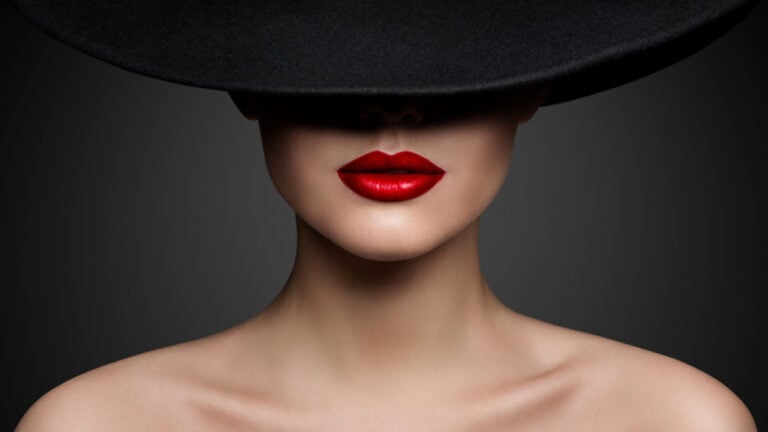 Red Lips Make up Closeup. Mysterious Fashion Woman Face Hidden by Black brimmed Hat. Elegant Retro Lady Fine Art Portrait over Gray Background.