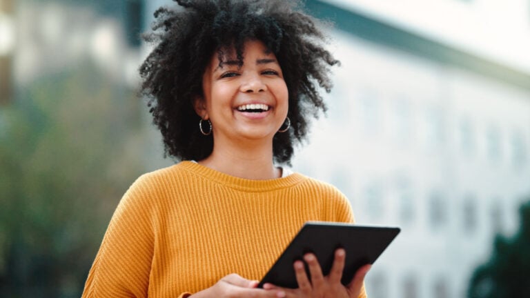 Portrait of happy woman with a digital tablet standing outside against a blurred urban background.