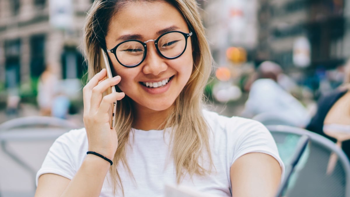 Cheerful asian girl enjoying communication with friend discussing plans while sitting in cafe in downtown,happy female student using connection in roaming for making calls abroad with cheap tariffs.