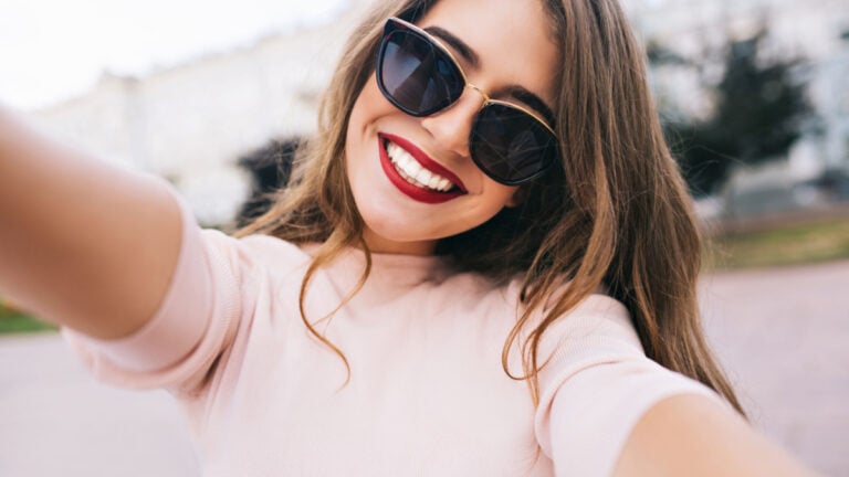 Closeup selfie-portrait student of attractive girl in sunglasses with long hairstyle and snow-white smile in city.