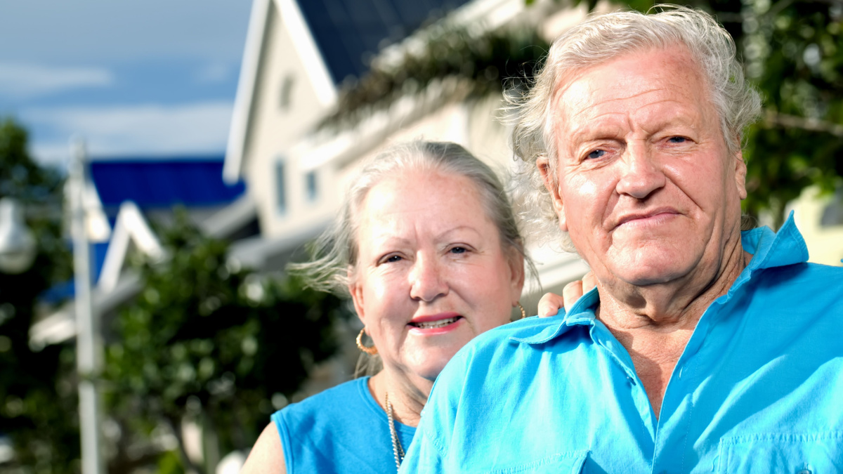A headshot of a happy elderly couple standing in front of a house.