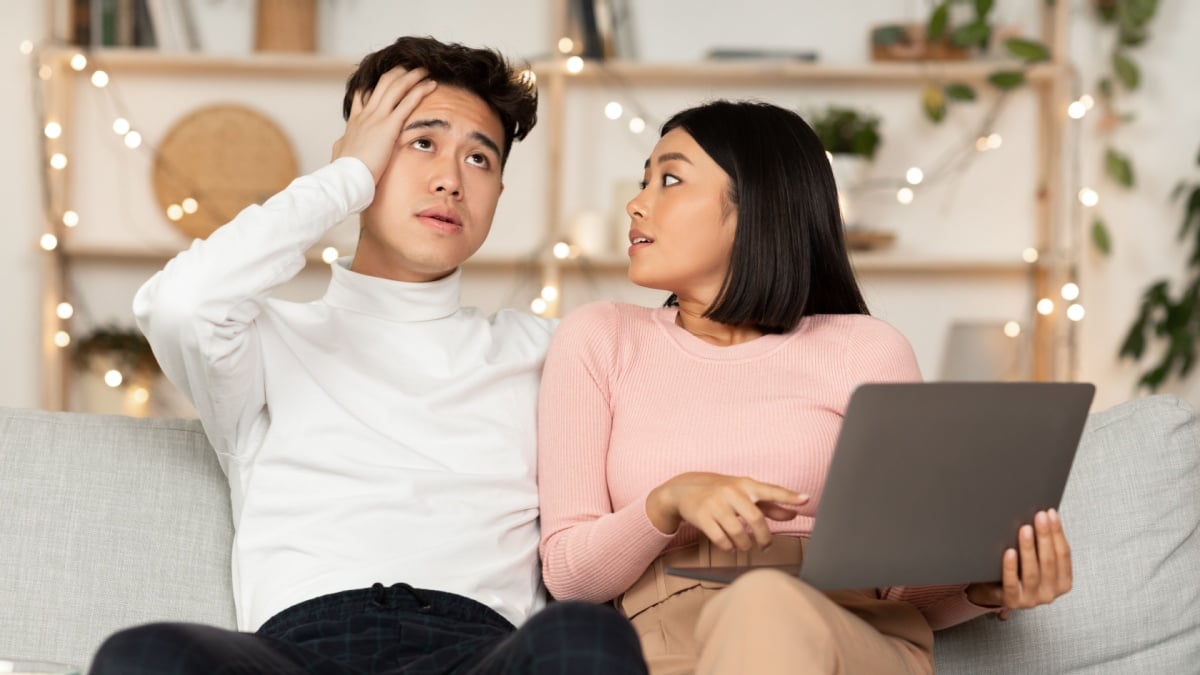 Asian Shopaholic Wife Shopping Online With Shocked Husband, Asking Him Buy Expensive Clothes And Spend Too Much Money Sitting On Sofa At Home.