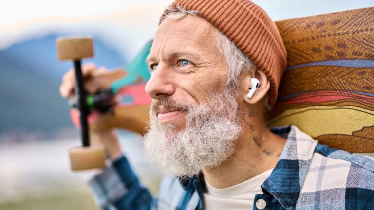 Active cool bearded old hipster man standing in nature park holding skateboard wearing earbud. Mature traveler skater enjoying freedom spirit and extreme sports hobby listening music in earphones.