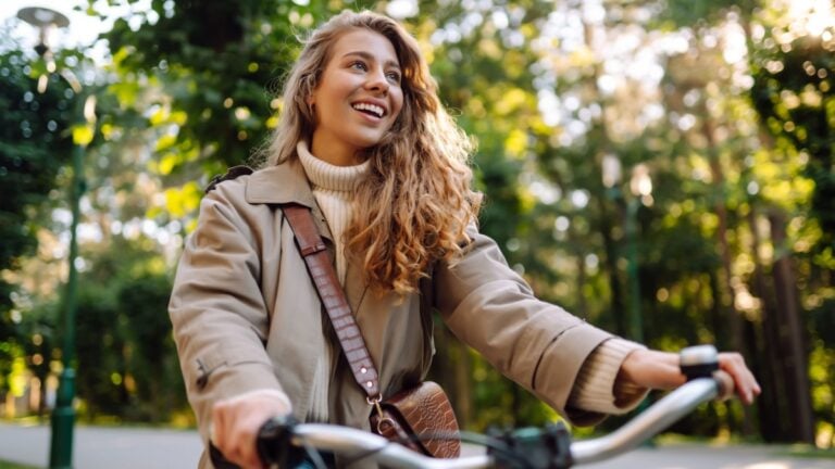 Smiling woman with curly hair in a coat rides a bicycle in a sunny park.