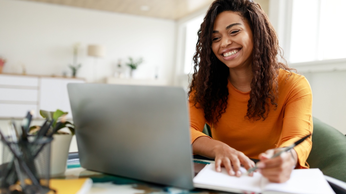 Portrait of smiling woman sitting at desk, using laptop and writing in notebook, taking notes, watching tutorial, lecture or webinar, studying online at home looking at screen.