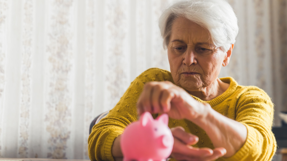 Medium indoor shot of an elderly short-haired woman in a yellow sweater putting her coins into the pink piggy bank.