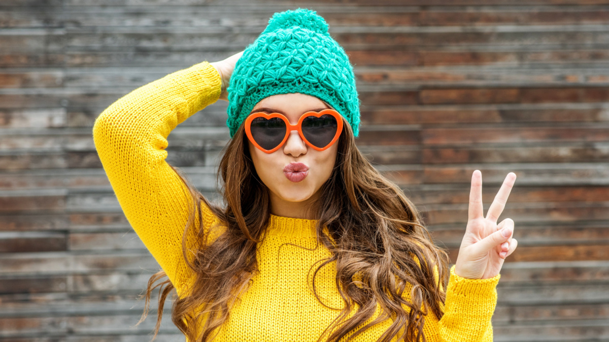 Beautiful brunette woman in sunglasses and knitted cap blowing lips kiss over wooden background.