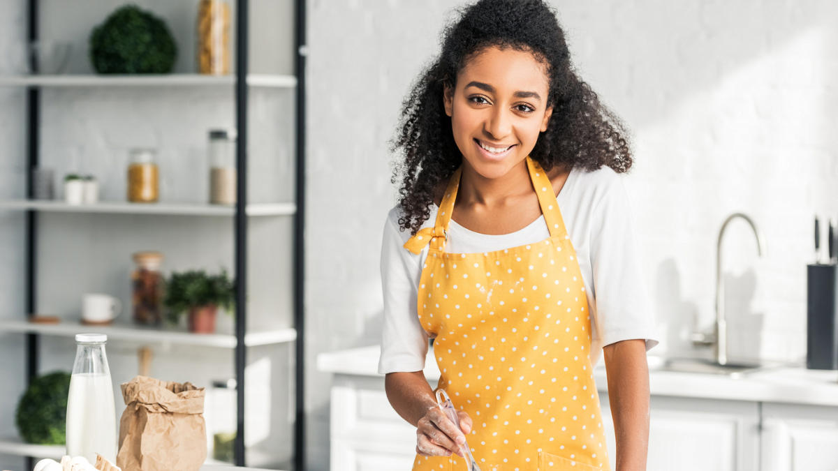 Beautiful smiling african american girl in apron applying oil on unbaked cookies in kitchen and looking at camera.
