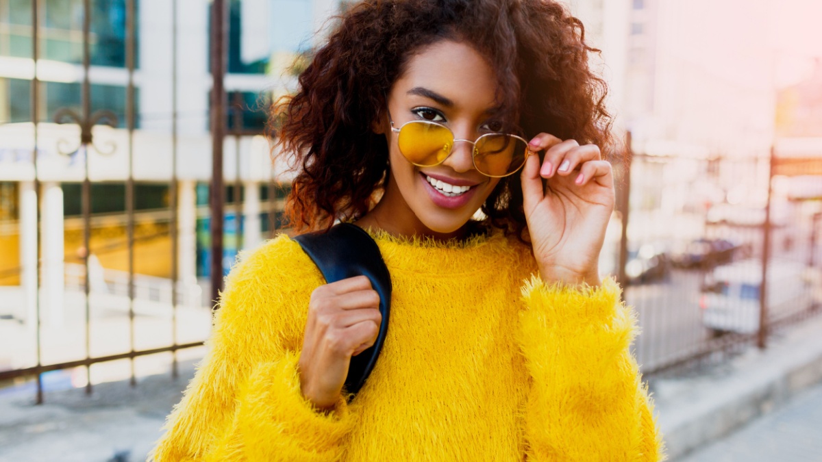 Close up portrait of smiling black woman in the stylist yellow sweater and sunglasses enjoying sunny day. College girl walking over the city.